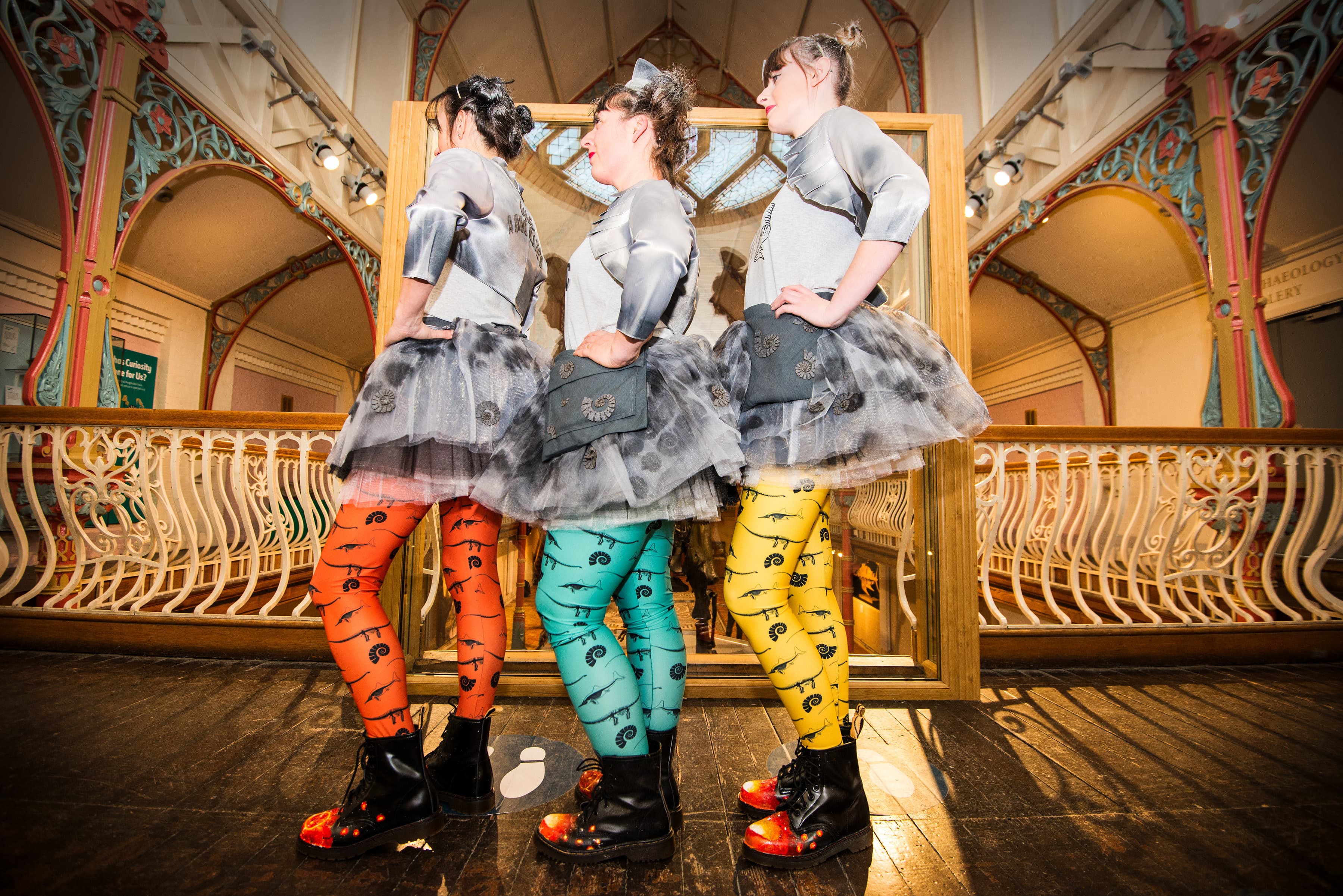 An image of 3 performers standing in a light facing the right wearing brighly colours leggings, large black boots and a grey skirt and bodic costume. They are standing infront of a wooden railed banister in an old style building.