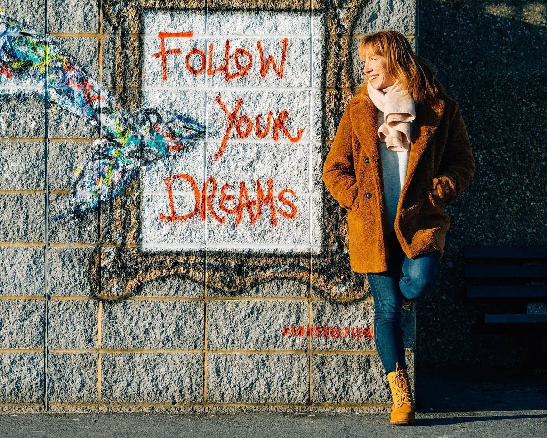 Photograph of Jane White standing name to text written on a wall that reads: Follow your dreams